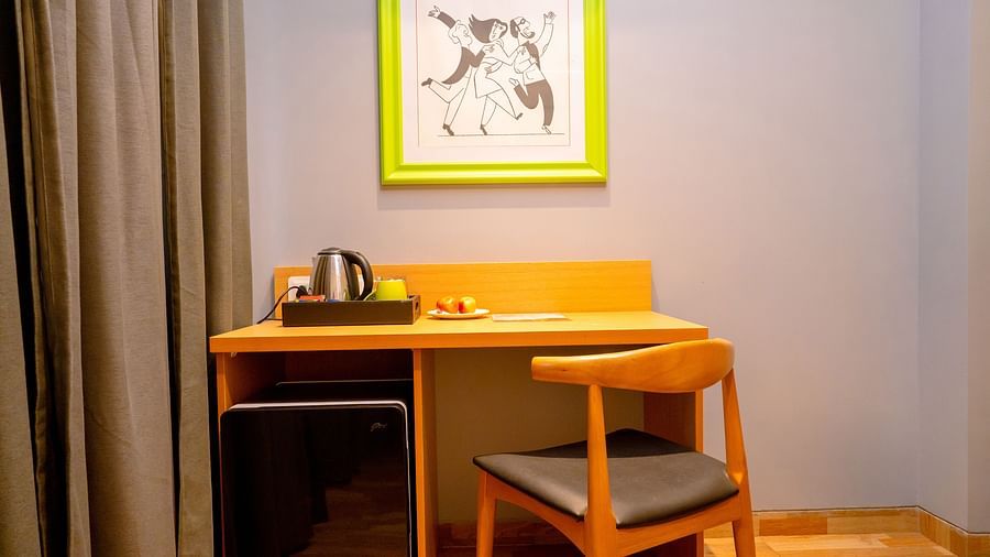 A cosy hotel room workspace with a wooden desk, chair and colorful artwork on the wall - TGI Ameya Grand, Goa