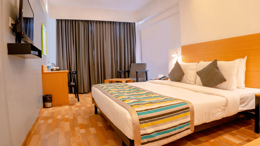 A bedroom at TGI Ameya Grand, Panjim, Goa, featuring a bed with a colourful runner, a solid headboard, a flat-screen TV, and a seating area. 