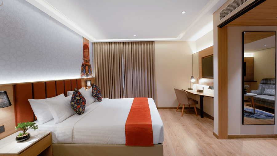 a modern hotel room at TGI Oberon featuring a large bed with white linens and red accents, padded headboard, pendant lights, wooden flooring, work desk with chair, wall?mounted television, and a curtain covering the window, creating a cozy and inviting atm