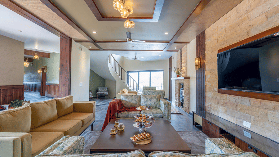 Ground floor lounge with beige and floral sofas, a stone-clad wall with television and fireplace, a glass pendant light and staircase at The Chamomile Estate.