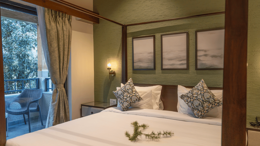 Oolong Room with a 4 poster bed in white linen, framed prints on a green wall and a glazed door to a balcony with wicker chairs at The Chamomile Estate.