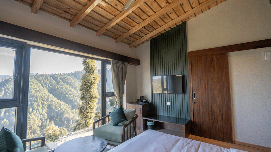 Sage Room seating with green armchairs, a round table, wall-mounted television, wardrobe and tea tray below a timber ceiling at The Chamomile Estate.