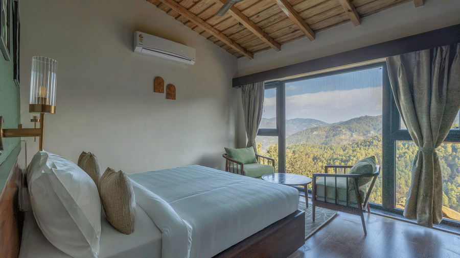 Sage Room with a bed in white linen, a brass wall light, green armchairs and a side table at a corner window over forested hills at The Chamomile Estate.