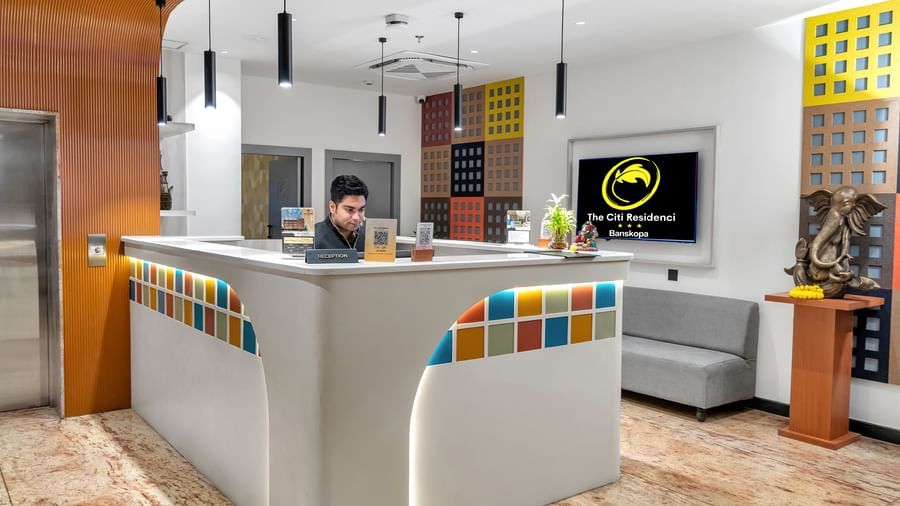 A hotel reception desk with colourful tiles and a staff member, in a lobby with a sofa and artwork | The Citi Residenci Hotel Banskopa
