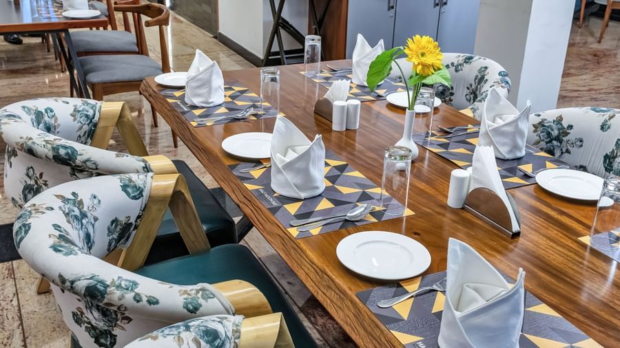 A long wooden dining table set with plates, folded napkins, glasses, and a yellow flower centerpiece, surrounded by chairs with floral patterned upholstery | The Citi Residenci Hotel Banskopa