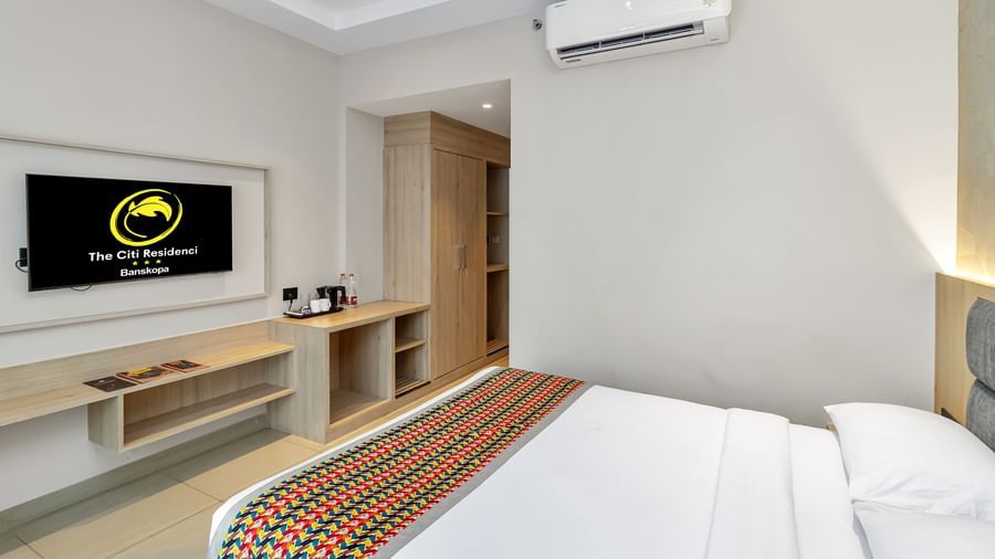 A  room with a bed featuring a colourful patterned runner, a wall-mounted TV displaying a logo, and an air conditioning unit  | The Citi Residenci Hotel Banskopa