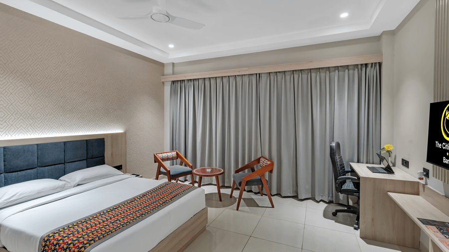 A well-lit hotel room with a queen size bed featuring a colourful patterned throw, a seating area with two chairs and a small table, a work desk with a chair, and a television | The Citi Residenci Hotel Banskopa