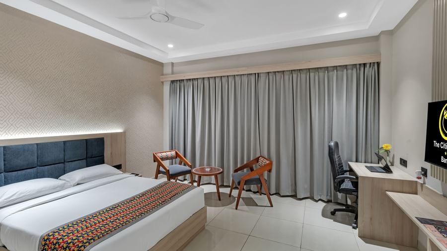 A well-lit hotel room with a king size bed featuring a colourful patterned throw, a seating area with two chairs and a small table, a work desk with a chair, and a television | The Citi Residenci Hotel Banskopa