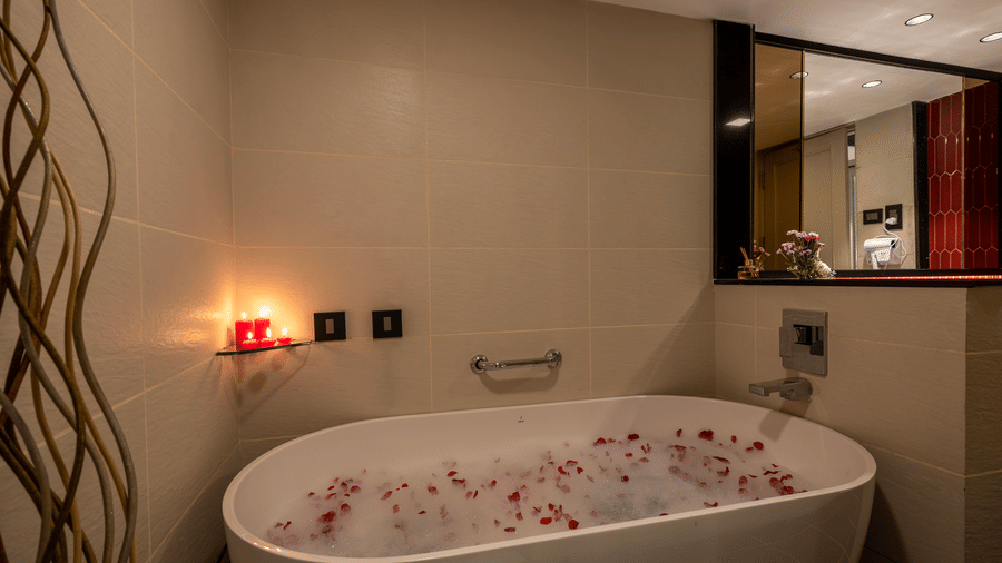 Close-up of a deep white soaking tub filled with water and rose petals, set against a warm-toned tiled wall with a lit candle in the background at The Citrine, Bangalore.
