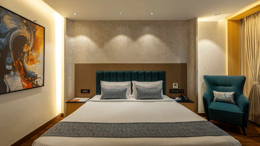 An executive king room featuring a large bed with a teal headboard, a blue armchair, and warm recessed lighting at The Citrine, Bangalore.