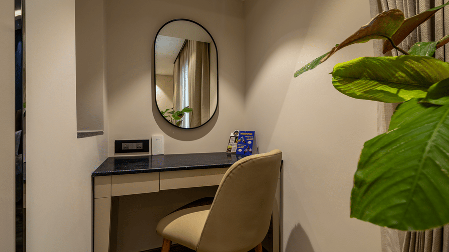 A dark wooden writing desk with a beige chair and an oval wall mirror next to a green plant at The Citrine, Bangalore.