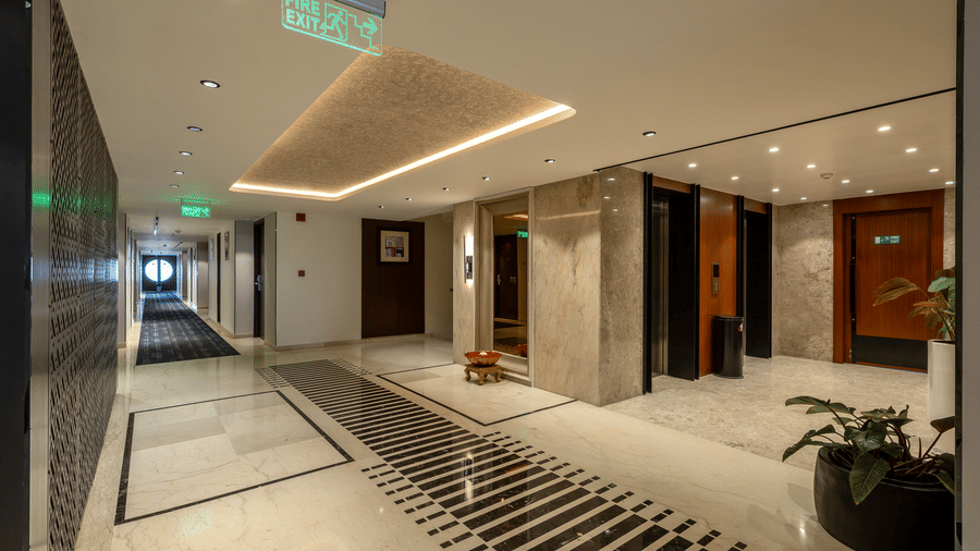 Lift lobby corridor at The Citrine, Bangalore, with marble flooring, illuminated fire exit sign, recessed ceiling lights, and lift doors on the right.