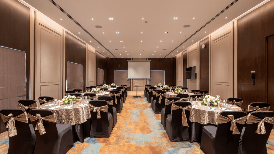 Spacious hotel conference room set for a banquet with long rectangular tables, dark chairs, patterned carpet, and a projection screen at the far end at The Citrine, Bangalore.