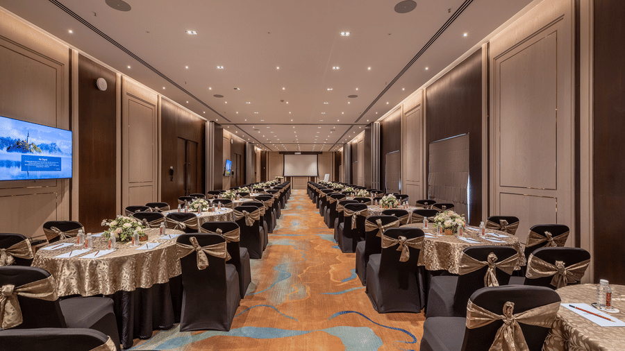 Wide hotel conference room arranged for a dining event with rows of round tables, dark chairs, a projection screen on the left, and warm overhead lighting at The Citrine, Bangalore.