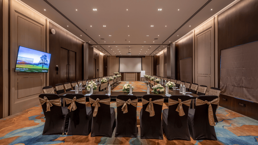 Large, modern conference room at Nexus set up with round tables, black chairs with gold bows, and a projector screen at The Citrine, Bangalore.
