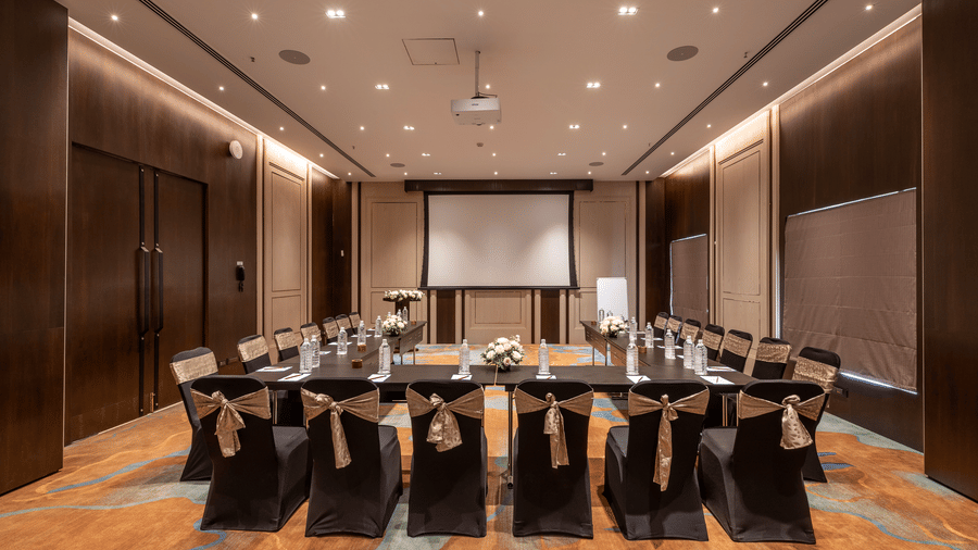Conference room at Nexus set up with round tables, black chairs with gold bows, and a projector screen at The Citrine, Bangalore.