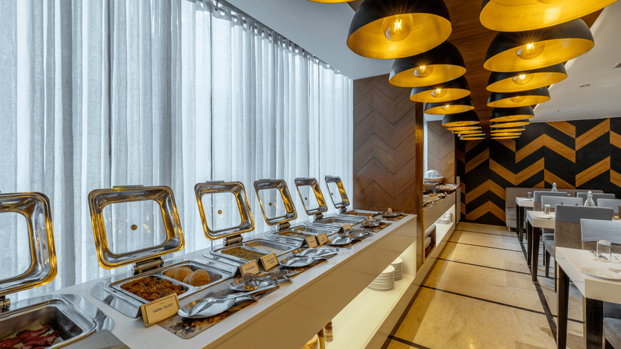 Buffet line at the restaurant of The Citrine, Bangalore, with stainless steel chafing dishes, sheer curtains, and black-and-gold pendant lamps overhead.