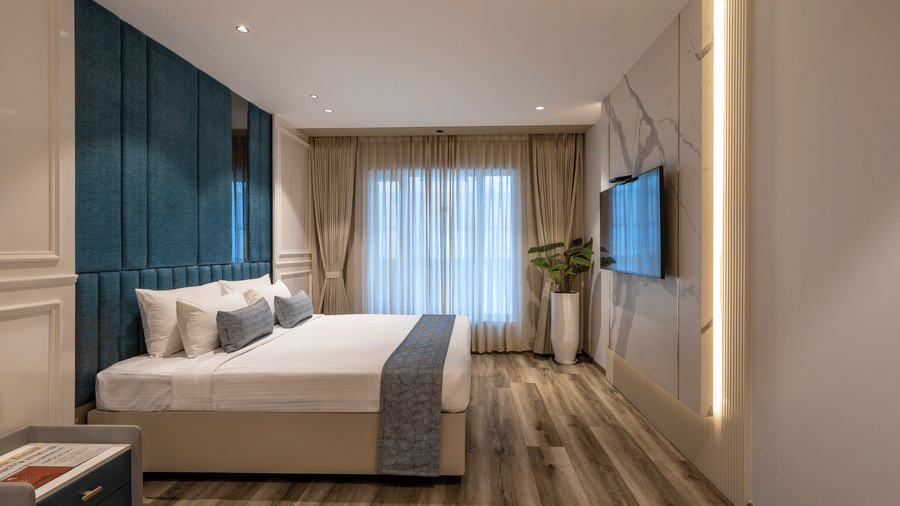 Suite bedroom at The Citrine, Bangalore, with a king bed, blue tufted upholstered headboard, wall-mounted TV on marble panel, and sheer curtains.