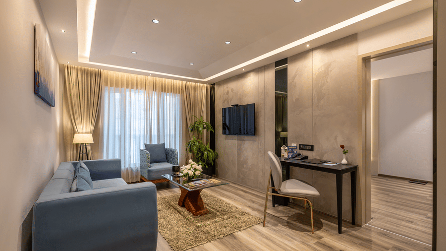 Suite living area at The Citrine, Bangalore, with a blue sofa, accent chair, wall-mounted TV, work desk, and beige curtains over a window.