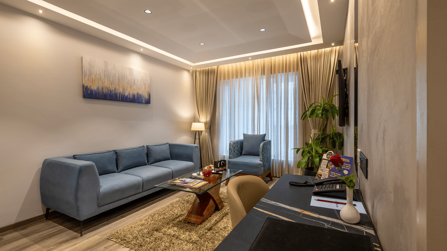 Suite lounge at The Citrine, Bangalore, with a blue sofa, armchair, artwork above the couch, floor lamp, and a black work desk in the corner.