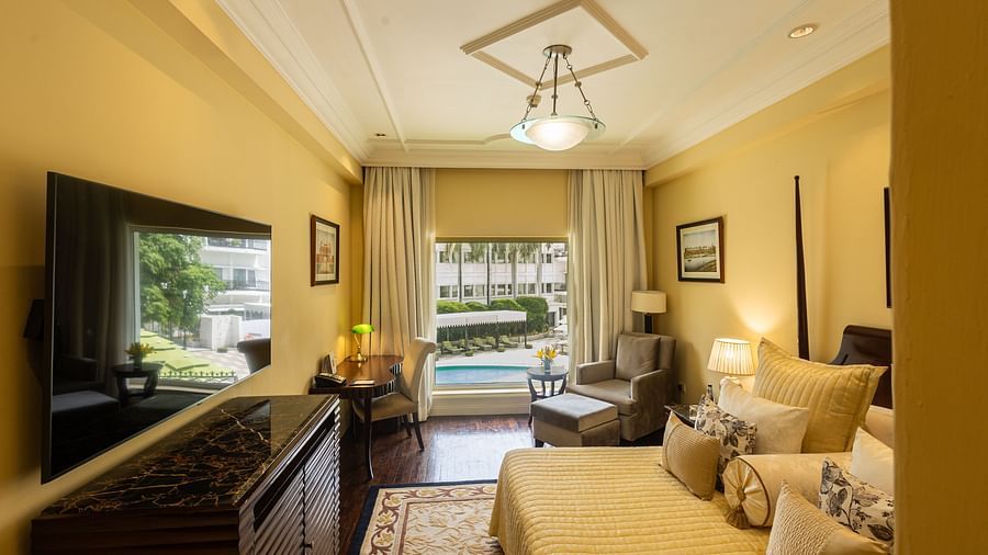 Side view of the Deluxe King Bed Room Pool View at The Claridges New Delhi, featuring a cosy king sized bed, wall-mounted TV, a corner chair, and a large window overlooking the swimming pool