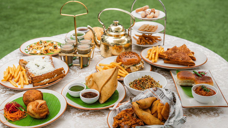 A breakfast table where varieties of savouries served in each plat consisting of samosa, french fries, sandwiches, salsa, fritters, bun, etc. There is also a kettle and hot tea served in four cups. 
