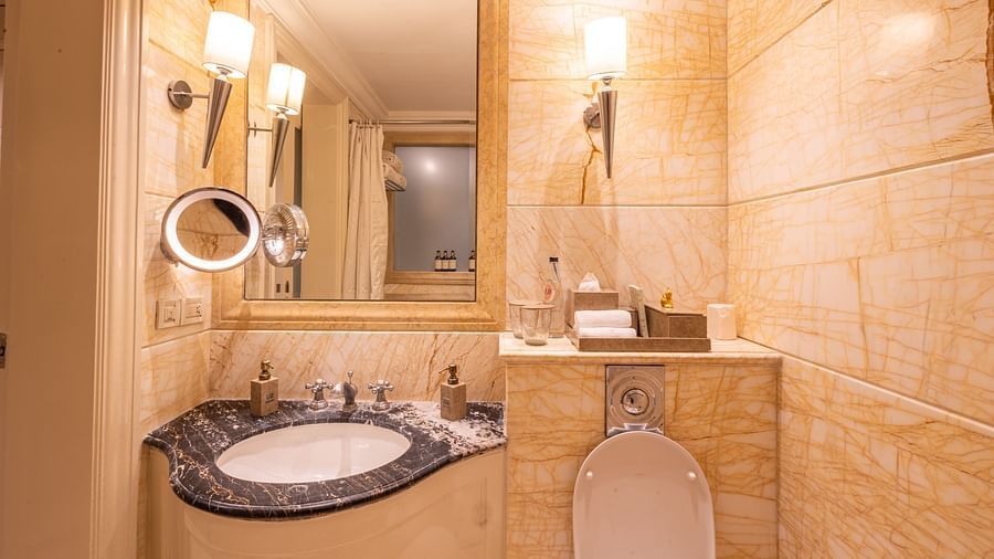 image of a bathroom with a small sink and a square mirror above it and a commode besides it at out heritage parlour rooms -  The Claridges New Delhi