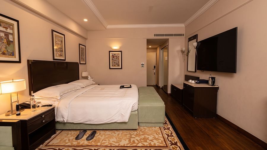 image of a brightly lit room with a king size bed and a wall mounted television placed opposite to the bed at our heritage balcony room - image of the heritage balcony room with sleek interiors and a a plush king size bed - The Claridges New Delhi