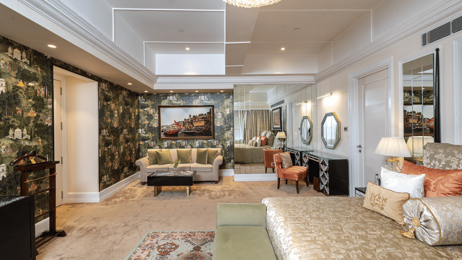 Presidential Suite bedroom featuring a king size bed, bedside tables, television, upholstered seating, and a large patterned area rug at The Claridges New Delhi.