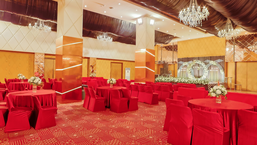 A section of a banquet hall featuring round tables draped in red, with floral centrepieces at The Conway, Meerut.