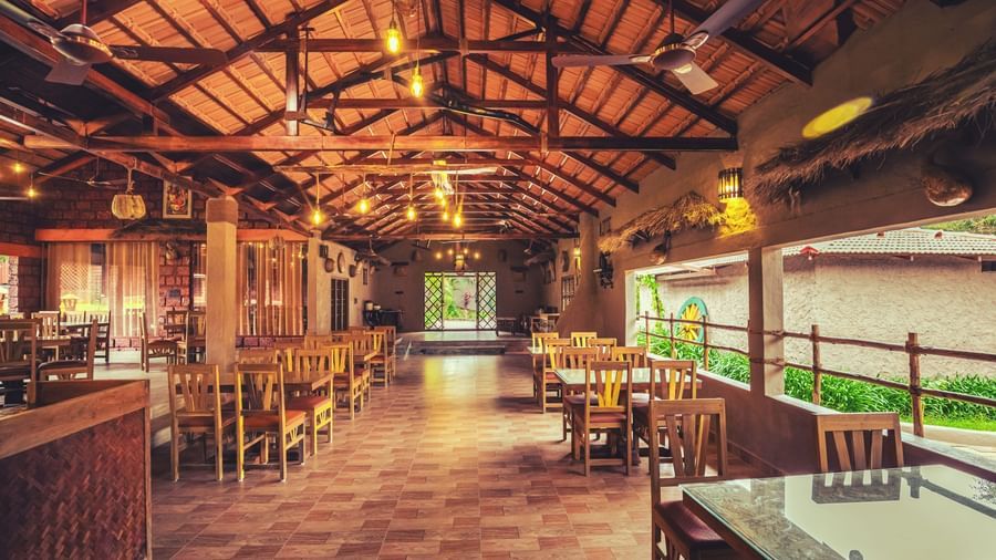 An interior shot of a spacious, open-air restaurant with a high wooden ceiling, many tables and chairs, and a view of the outdoors at The Copper Edge - Eco Resort, Coorg.