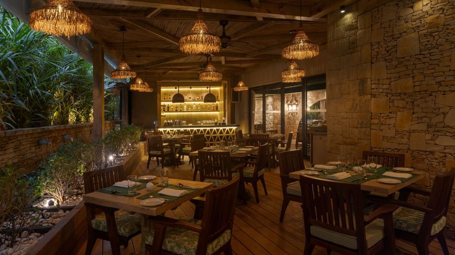 A dimly-lit dining area at the Mila Restaurant with lush green trees on one side at The Evren, Vagator.
