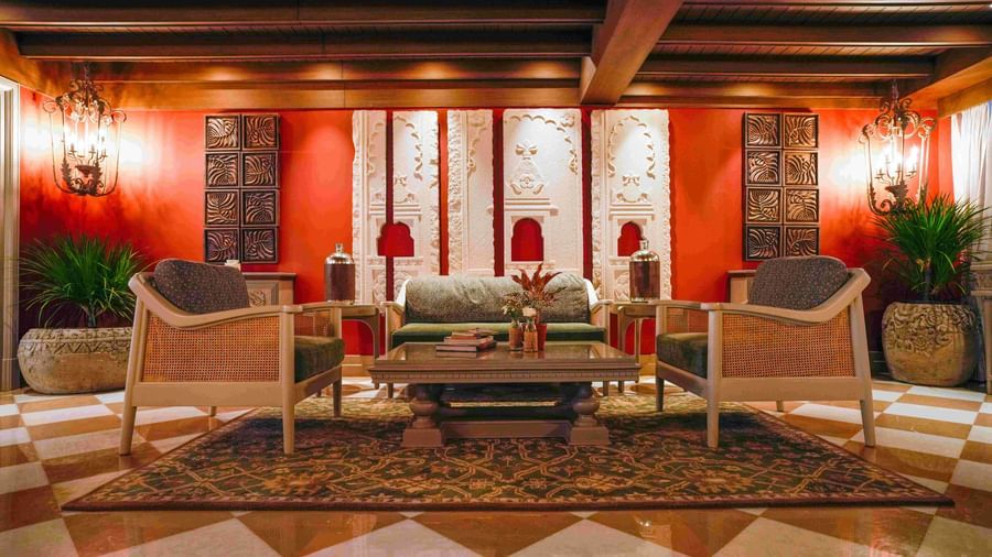 A wide shot of a welcoming lounge area at The Evren, Vagator. The space features a green sofa and two chairs with cane details, set against a vibrant red wall with ornate white carvings. The room has a checkered floor and a decorative rug.