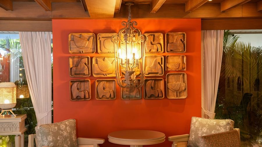 A cozy sitting area at The Evren, Vagator, with two chairs and a round table. The striking red wall is decorated with a series of carved wooden panels and illuminated by an elaborate chandelier.