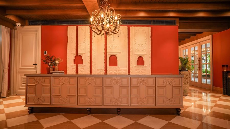 The reception or check-in area at The Evren, Vagator, featuring a large, ornate desk with detailed paneling. The background wall is a bold red with intricate white carvings, and a beautiful chandelier hangs from the ceiling.