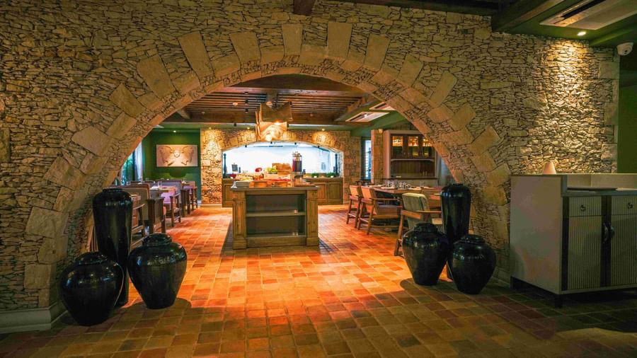 A view into Mila Restaurant at The Evren, Vagator, taken through a rustic stone archway. The restaurant has a tiled floor, a central buffet area, and various dining tables, creating a warm and inviting atmosphere.