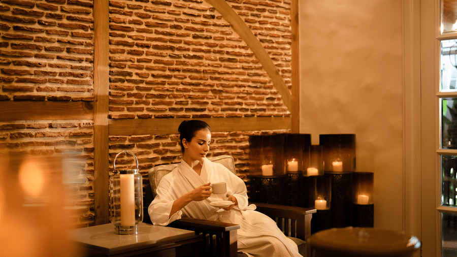A woman seated on a cushioned bench wearing a robe, with candles placed on a low table and textured brick walls behind at The Evren, Vagator.