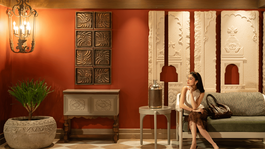 A woman seated on a chair in an indoor seating area with framed wall panels, table lamps, floor tiles, and potted plants at The Evren, Vagator.