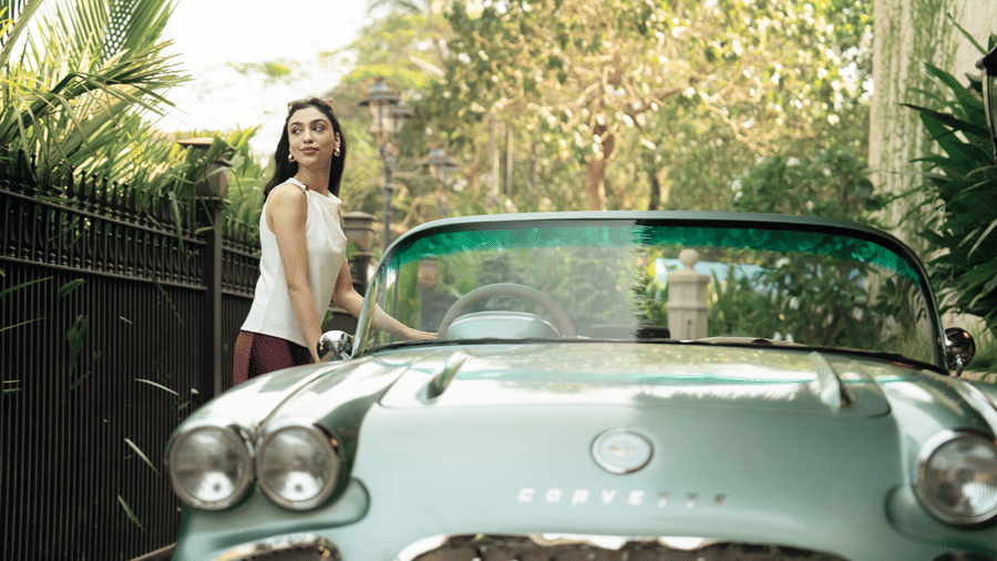 A woman standing beside a light colored vintage car with round headlights and a garden pathway behind at The Evren, Vagator.