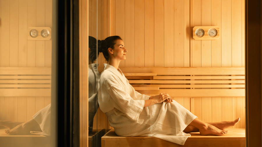 A woman seated inside a wooden sauna room wearing a robe, with slatted wooden walls and a glass door visible at The Evren, Vagator.