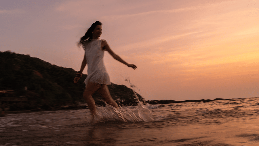 A woman walking through shallow seawater with waves around her feet, with a rocky shoreline and sunset sky visible at The Evren, Vagator.