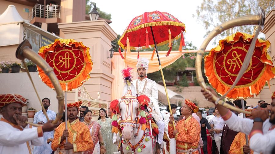 A traditional Indian wedding procession with people in orange carrying ornate umbrellas and flags, accompanied by music - The Grand Legacy Resort and Spa