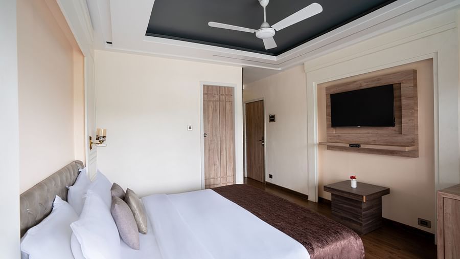 A spacious room with neutral-toned walls, a large bed, a flat-screen TV, and large windows offering natural light.