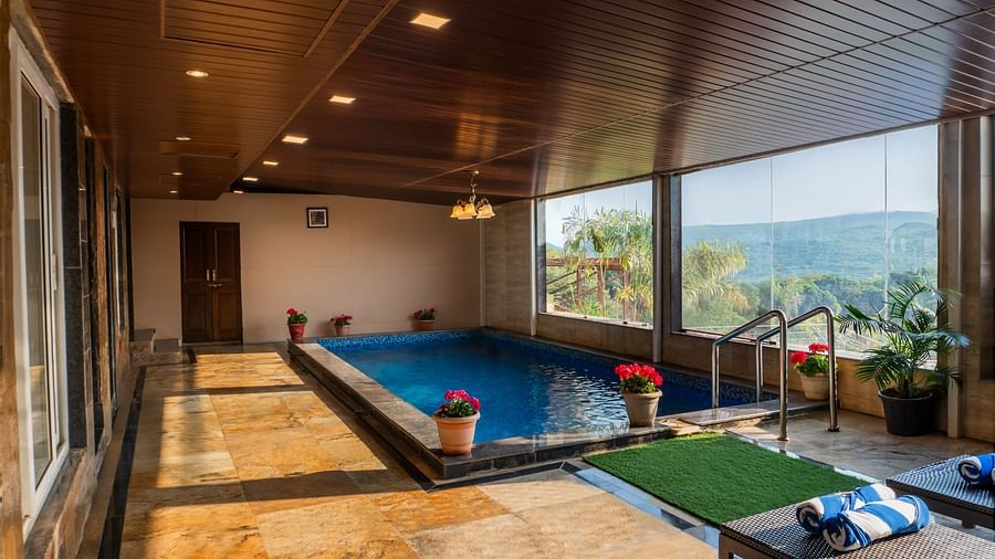 A bright indoor pool reflects the wood ceiling and recessed lights. Large windows reveal a scenic outdoor view. Lounge chairs with blue striped towels sit poolside on a stone floor next to potted flowers and a palm - The Grand Legacy Resort and Spa