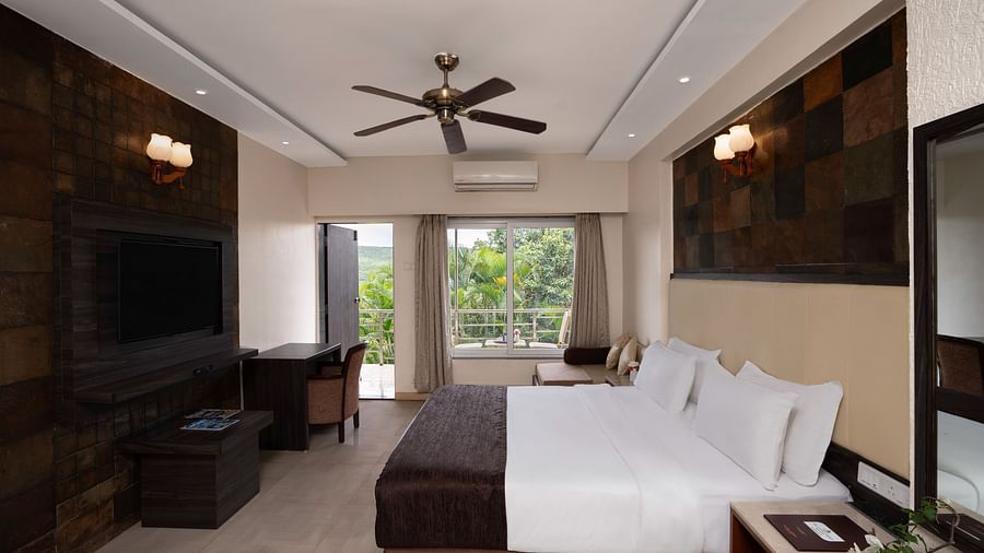 A spacious room with neutral-toned walls, a large bed, a flat-screen TV, and large windows offering natural light.