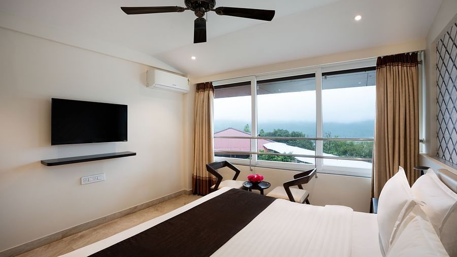 A spacious room with neutral-toned walls, a large bed, a flat-screen TV, and large windows offering natural light.
