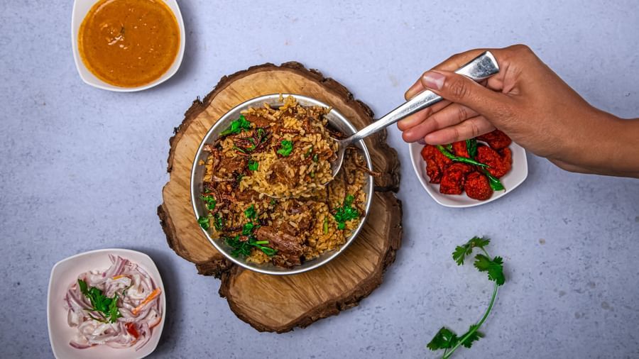A hand scooping from a bowl of richly garnished Indian curry served on a wooden platter, accompanied by bowls of chutney placed on a light blue surface at The Hamuse Hotel, Kodaikanal.