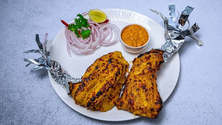 A plate of grilled tandoori chicken legs wrapped in foil at the ends, served with sliced onions, a lemon wedge on a white background at The Hamuse Hotel, Kodaikanal.
