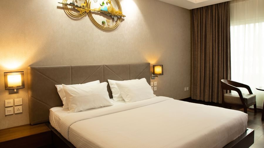 A bedroom with a large bed, bedside lamps, curtains drawn to the side, and natural light coming through the window at The Hamuse Hotel, Kodaikanal.