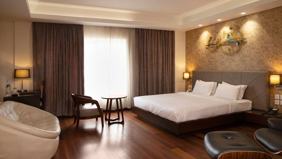 A spacious room featuring a large bed, a cosy seating area, elegant curtains, soft ambient lighting, and expansive windows with curtains drawn to the side at The Hamuse Hotel, Kodaikanal.